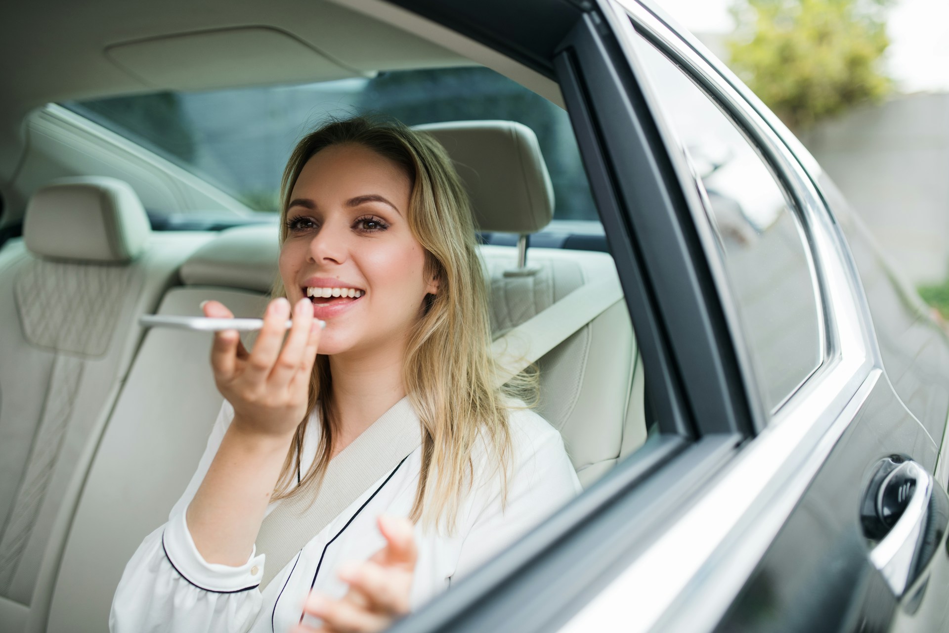 A happy woman in the back seat of a car holds a smartphone and records a voice message.